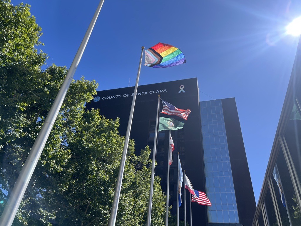 a picture of flags, including the pride flag and the American Flag lined in a row in front of trees and a building that says county of santa clara 