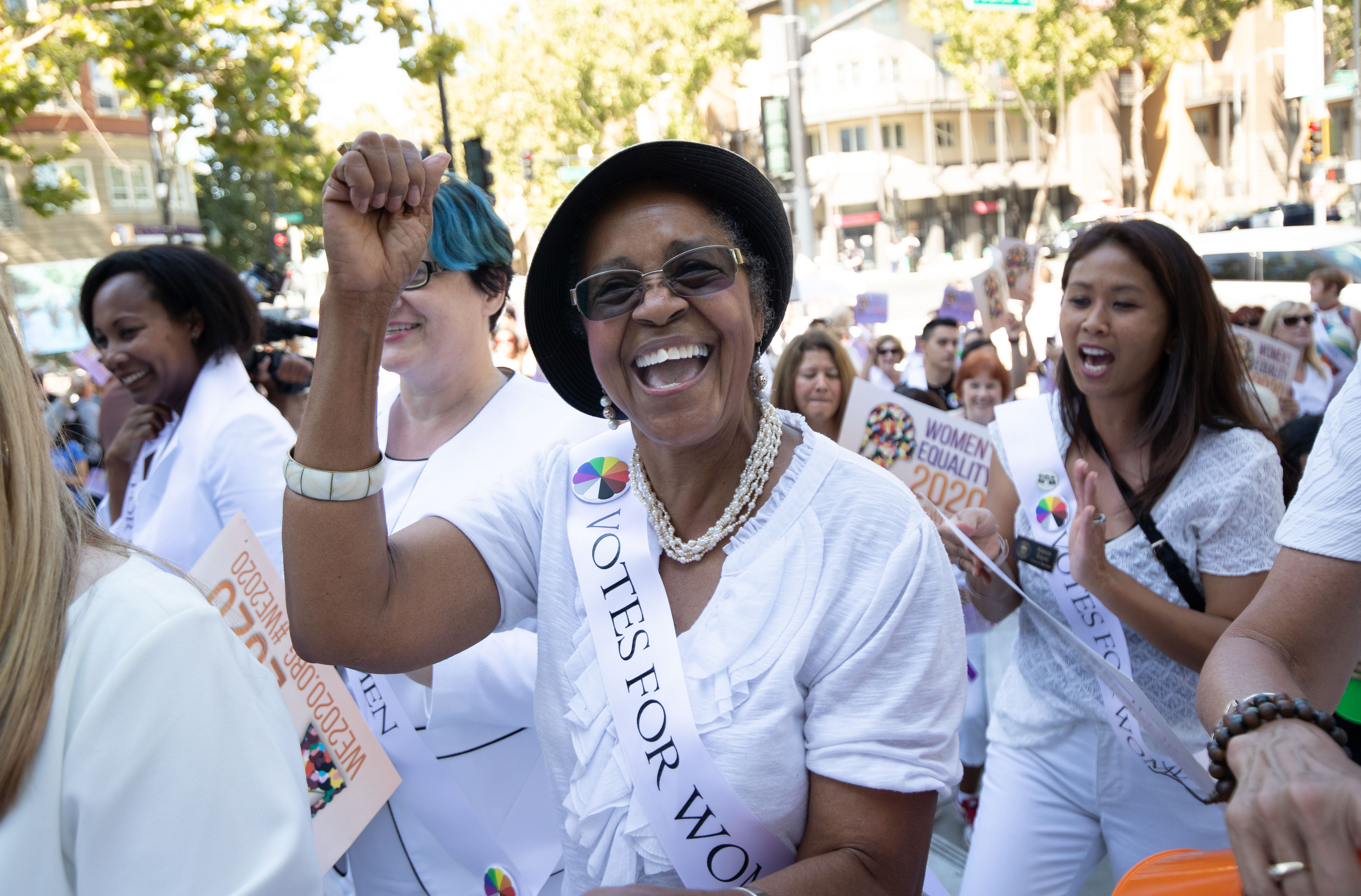 Person smiling in crowd with arm in the air, wearing a sash that reads "Votes for Women", at rally celebrating the 100th anniversary of women's right to vote, diverse crowd