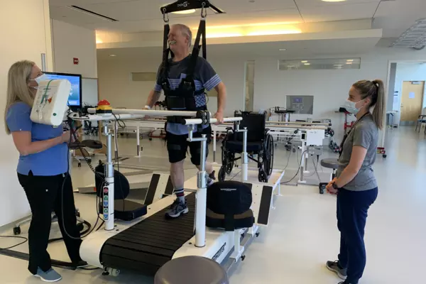 Two staff members at the Rehabilitation Center at VMC assist a patient in the use of a gravity harness, which supports him as he walks on a treadmill.