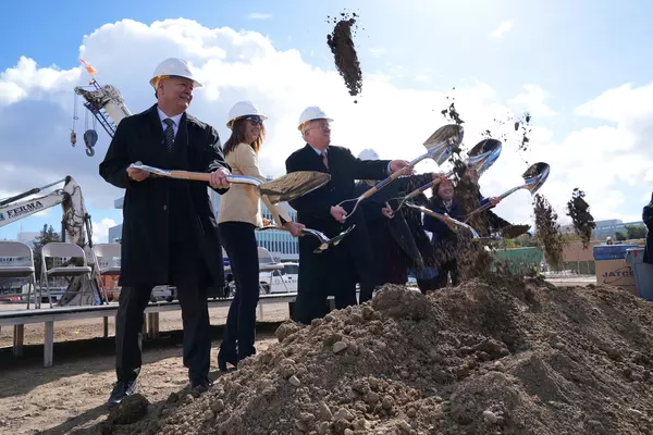 County of Santa Clara officials wearing hardhats break ground at the site of the future mental health services center on the Valley Medical Center campus in San Jose. They are standing in a line behind a pile of dirt, each with a shovel in hand, smiling and tossing dirt for the ceremony.