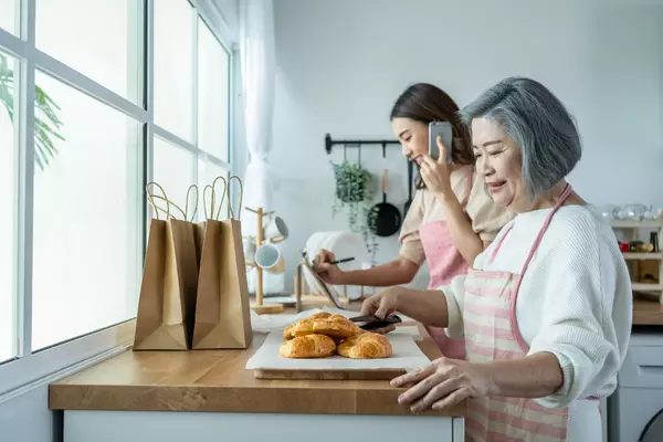 A bakery team -- possibly mother and daughter -- have a tray of croissants that they are putting in brown bags, while the younger woman is on the phone taking an order.