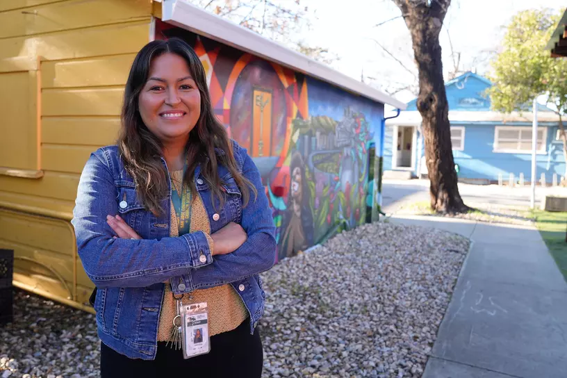 Natali Zepeda stands in front of colorful building, smiling with her arms crossed