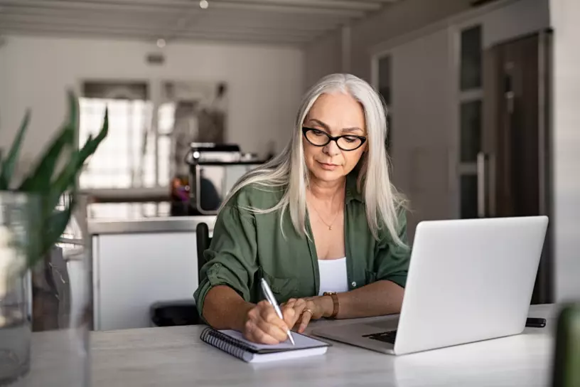 Middle-aged white woman wearing glasses and taking notes in front of a laptop