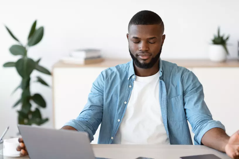 Black man closing eyes and relaxing at desk in front of a laptop