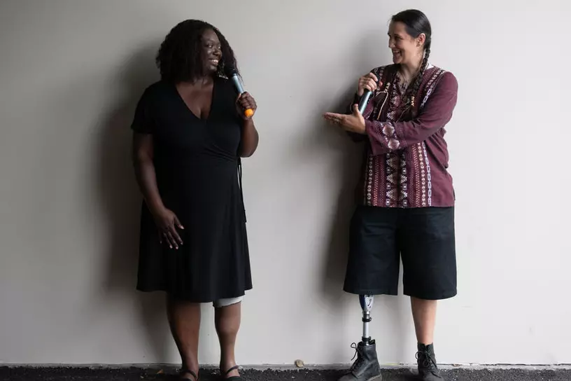 An Indigenous Two-Spirit person and a Black woman face each other and gesture while smiling and speaking into microphones. They are both wearing prosthetic legs.