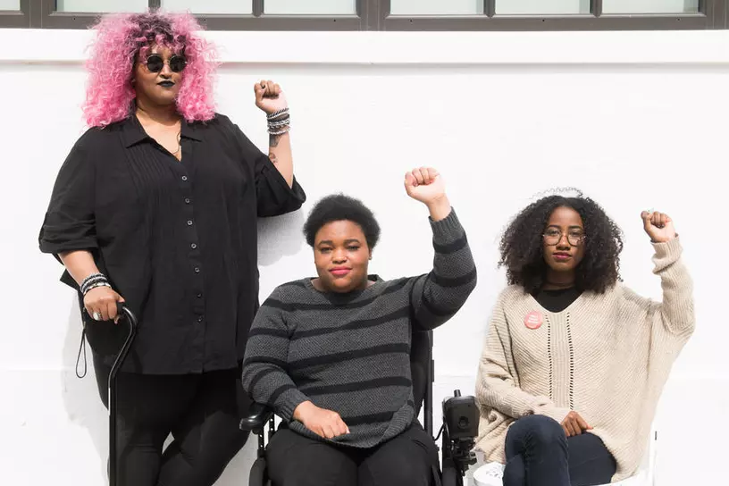 Three Black and disabled folx raise fists in front of a white wall. On the left, a non-binary person raises her right fist while holding her cane. On the right, a non-binary person in a power wheelchair and a femme in a folding chair have their left fists up.