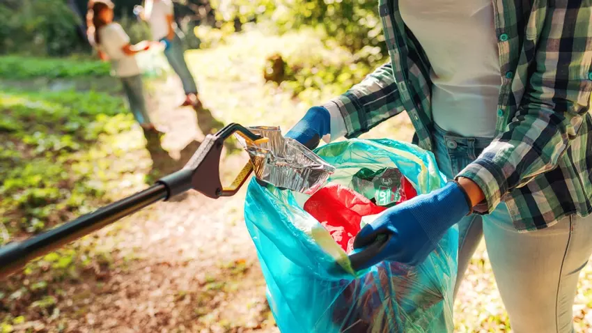 A person holding a garbage bag open while someone else puts a piece of trash in the bag using a litter grabber