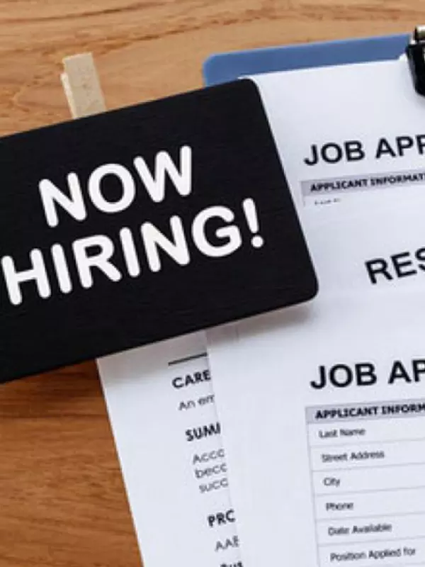 A clip-on badge with the text "Now Hiring!" sits on top of a stack of job application forms.