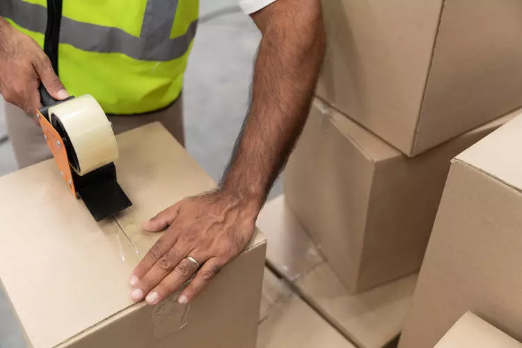 A person with a wedding band on their ring finger taping up a package with a pile of other packages on their left.
