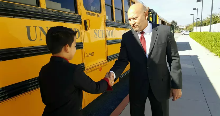 Community Prosecutor Johnny Gogo greets a student with a handshake next to a yellow school bus.