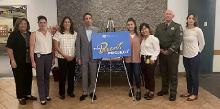 8 people, including District Attorney Jeff Rosen, standing in front of a blue sign that is on an easel that says "Parent Project"